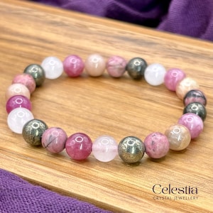 May include: A bracelet made of round beads in shades of pink, white, and metallic colours. The beads are strung together to form a circular bracelet. The bracelet is displayed on a wooden surface. The brand name "Celestia" is visible.