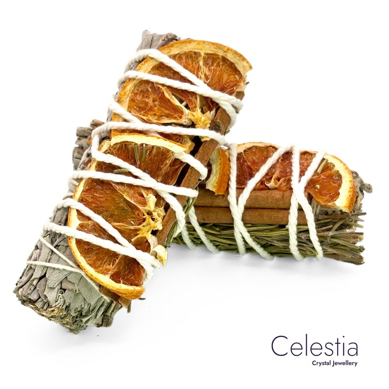 May include: Two sage bundles with dried orange slices, cinnamon sticks, and rosemary, wrapped with white twine. The bundles are arranged on a white background. The text "Celestia Crystal Jewellery" is in the bottom right corner.