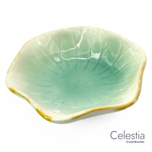 May include: A small, ceramic dish with a unique, petal-like shape. The dish features a gradient of colors, transitioning from a light green center to a darker green hue, with a cream-colored rim and a gold-colored edge. The text "Celestia Crystal Bracelets" is visible.