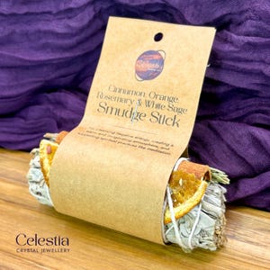 May include: A smudge stick bundle with a brown paper label. The label reads "Cinnamon, Orange, Rosemary & White Sage Smudge Stick." The bundle includes dried orange slices, sage, and rosemary, tied with twine. The Celestia Crystal Jewellery logo is at the bottom.