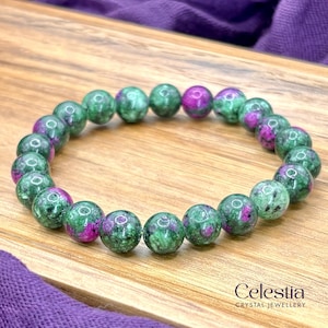May include: A beaded bracelet featuring green and magenta gemstones. The round beads are a mix of green with magenta inclusions. The bracelet is displayed on a wooden surface. The brand name "Celestia Crystal Jewellery" is visible.
