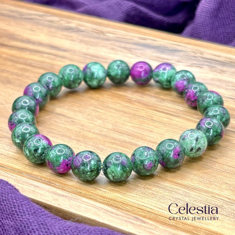 May include: A beaded bracelet featuring green and magenta gemstones. The round beads are a mix of green with magenta inclusions. The bracelet is displayed on a wooden surface. The brand name "Celestia Crystal Jewellery" is visible.