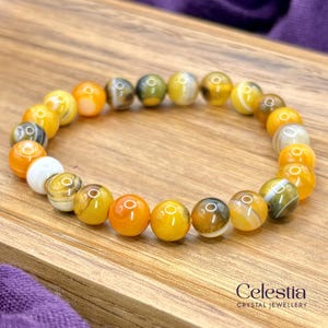 May include: A bracelet made of round, polished beads in shades of yellow, orange, white, and grey. The beads are strung together to form a bracelet. The bracelet is displayed on a wooden surface. The Celestia Crystal Jewellery logo is visible.