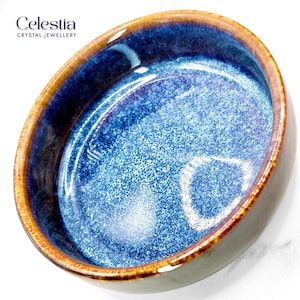 May include: A round ceramic bowl featuring a deep blue interior with a speckled texture and a smooth, glossy finish. The rim is accented with a warm, golden-brown glaze. The text Celestia CRYSTAL JEWELLERY is printed in the upper left corner.