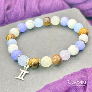 Puede incluir: Una pulsera de cuentas con piedras preciosas coloridas, incluyendo ojo de tigre, cuentas azules, blancas y amarillo claro. Un dije plateado con el símbolo de Géminis cuelga de la pulsera. El logo de Celestia Crystal Jewellery es visible.