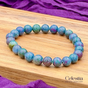 May include: A stretch bracelet composed of round, polished beads in shades of blue, green, and magenta. The beads feature a mottled, natural stone texture with varying colour intensity throughout each piece. The text Celestia Crystal Jewellery is visible.