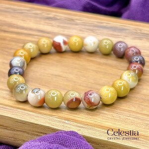 May include: A bracelet made of round, multi-coloured gemstones, predominantly yellow with red, white, and grey accents. The bracelet is displayed on a wooden surface. The Celestia Crystal Jewellery logo is visible.