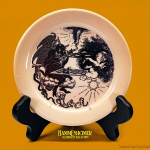 May include: A cream-colored decorative plate featuring a black and white illustration of a devil playing chess against an angel. The plate is displayed on a black stand against a yellow background. The text "HAMMERHEIMER A CURIOSITY COLLECTION" is at the bottom.
