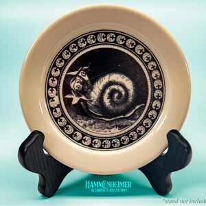 May include: A cream-colored decorative plate with a black and white illustration of a snail with a person riding it. The plate has a black border with cross-like designs. The plate is displayed on a black stand. The text "HAMMENHEIMER A CURIOSITY COLLECTION" is at the bottom.