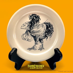 May include: A white ceramic plate featuring a whimsical black and white illustration of a chicken in a dress, holding a fan. The plate is displayed on a black stand against a yellow background. The text "HAMMENHEIMER A CURIOSITY COLLECTION" is at the bottom.