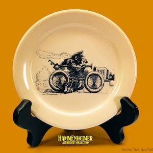 May include: Cream-colored decorative plate featuring a black and white illustration of a person driving a vintage car. The plate has a glossy finish and is displayed on a black stand. The plate has the text "HAMMENHEIMER" and "A CURIOSITY COLLECTION".