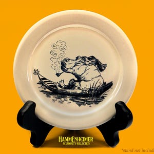 May include: A cream-colored decorative plate featuring a whimsical illustration of a hippo smoking a pipe while seated in a boat. The plate is supported by a black stand against a yellow background. The plate has the text "HAMMENHEIMER ACUROSITY COLLECTION".
