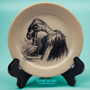 May include: A cream-colored decorative plate featuring a black and white illustration of a bird-like creature looking through a microscope. The plate is displayed on a black stand. The text "HAMMENHEIMER A CURIOSITY COLLECTION" is at the bottom.