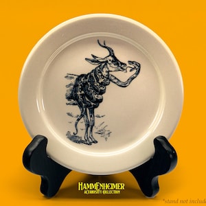 May include: A white ceramic plate with a black illustration of a deer-like creature holding a snake. The plate is displayed on a black stand against a yellow background. The text "HAMMENHEIMER ACURIOSITY COLLECTION" is at the bottom.