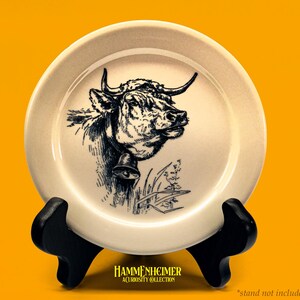 May include: Cream-colored decorative plate featuring a detailed black and white illustration of a cow's head with horns and a bell. The plate is displayed on a black stand against a bright yellow background. The text "HAMMENHEIMER A CURIOSITY COLLECTION" is at the bottom.