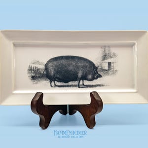 May include: Rectangular off-white ceramic serving platter with a black and white illustration of a pig. The platter is supported by a dark brown wooden stand. The platter has a slightly raised edge.