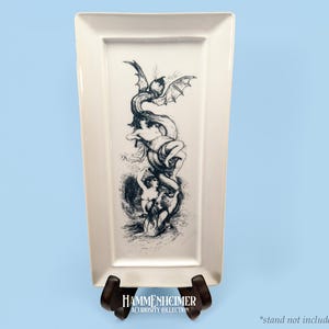 May include: Rectangular off-white ceramic serving tray with a black and white illustration of a dragon-like creature intertwined with figures. The tray is displayed on a dark brown stand against a light blue background. The text "HAMMENHEIMER A CURIOSITY COLLECTION" is visible.