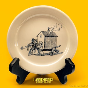 May include: A cream-colored plate with a whimsical black and white illustration of a house on a creature's back, with smoke rising from the chimney. The plate is displayed on a black stand against a yellow background. The text "HAMMENHEIMER ACURIOSITY COLLECTION" is at the bottom.