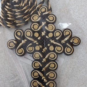 May include: A decorative cross-shaped embellishment with a braided cord. The cross is black with gold-colored spiral accents. The cord is black and gold. The item is in a clear plastic bag.