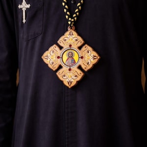 May include: A religious pendant featuring a central icon of a figure with a halo, set within a decorative, light-colored wooden cross. The cross is adorned with gold-colored embellishments and red accents. The pendant hangs from a black and gold braided cord.