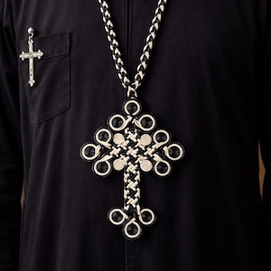 May include: A black and white woven cross pendant necklace. The cross is intricately designed with circular and woven elements. A small silver cross pin is on the left side of the black garment.