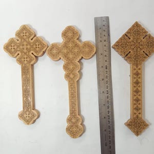 May include: Three wooden crosses with detailed carved patterns. The crosses are light brown. A ruler is placed next to the crosses, showing measurements in centimetres.