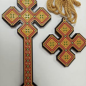 May include: Two wooden crosses with intricate geometric designs in red, yellow, and green. One is a large wall cross, the other a pendant. A gold-colored woven cord is included with the pendant.