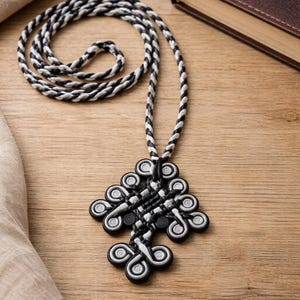 May include: A black and white pendant necklace with a braided cord. The pendant has a detailed, symmetrical design. A closed book with a gold cross design is in the background.