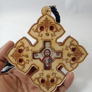 May include: Wooden Orthodox cross with ornate carvings, red jewel accents, and a central icon of Jesus Christ. The cross is suspended by a black cord and features a decorative design.