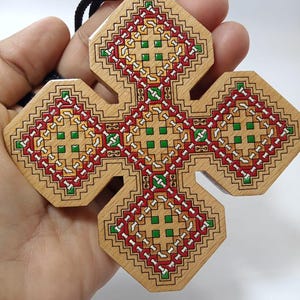 May include: Wooden cross pendant with a geometric design in red, green, yellow, and white. The cross is held by a black cord. The cross has a light brown wood grain.
