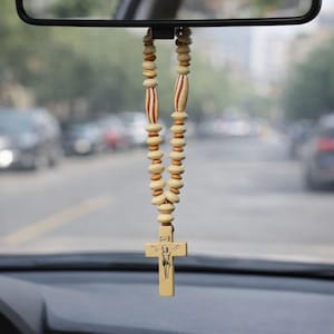 May include: A wooden rosary hanging from a car's rearview mirror. The rosary features light-colored wooden beads and a cross pendant with a religious figure. The background shows a blurred street scene.