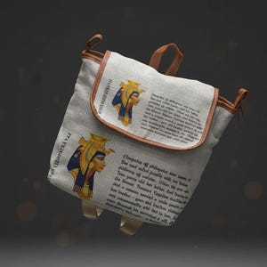 May include: A canvas backpack with brown leather trim and straps. The bag features illustrations of Cleopatra and text in a foreign language. The bag has a flap closure and gold-coloured hardware.
