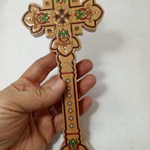 May include: Wooden cross with intricate carvings and colorful accents. The cross features a central design and decorative elements in red, green, and gold. The cross is held in a hand, showcasing its detailed craftsmanship.