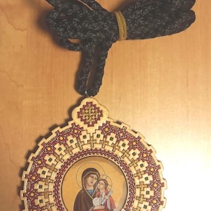 May include: Wooden religious pendant featuring an image of Mary and Jesus, surrounded by a decorative border with red, gold, and green accents. The pendant is suspended from a black and gray rope, secured with a yellow rubber band.