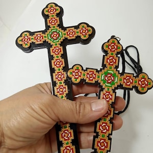 May include: Two decorative wooden crosses with intricate floral patterns in red, green, and gold against a black background. One cross has a black cord attached. The crosses are held in a hand against a white background.