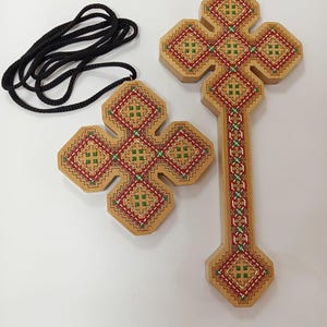 May include: Two wooden crosses with intricate red, green, and white embroidered patterns. One cross is larger with a long stem, while the other is smaller and attached to a black cord. The crosses are made of light-colored wood.