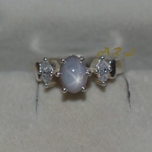 May include: A silver ring featuring a central oval, light blue star sapphire gemstone, flanked by two marquise-cut, clear gemstones. The ring has a simple, elegant design, perfect for everyday wear or special occasions.