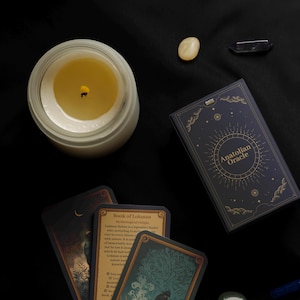 Peut inclure: Vue a&eacute;rienne d'une bougie allum&eacute;e dans un pot en verre, d'un jeu de cartes "Anatolian Oracle" et de plusieurs cristaux sur un tissu noir. Le jeu de cartes est bleu marine avec des lettres dor&eacute;es et un motif circulaire.