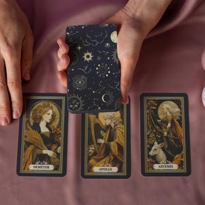 May include: A tarot card deck with a celestial design on the back is held above three face-up cards. The cards feature illustrations of Demeter, Apollo, and Artemis. Two pink candles sit to the right on a pink satin surface.