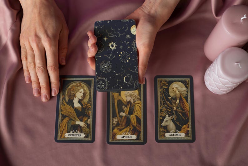 May include: A tarot card deck with a celestial design on the back is held above three face-up cards. The cards feature illustrations of Demeter, Apollo, and Artemis. Two pink candles sit to the right on a pink satin surface.