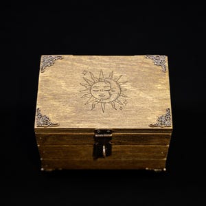 May include: A wooden box with a celestial design. The box is stained a warm brown color and features a laser-etched sun and moon design on the lid. Decorative metal corner accents and a clasp complete the design.