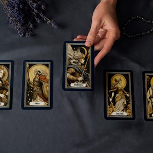 May include: A hand holding a tarot card with the name "Hermes" on it, surrounded by other tarot cards. The cards feature illustrations of figures with names like "Demeter", "Ares", "Themis", and "Zeus". The cards have a dark blue border.