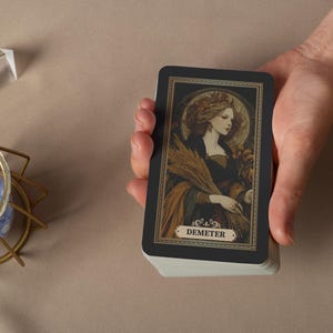 May include: A deck of tarot cards featuring a vintage illustration of Demeter, the Greek goddess of harvest. The card has a black border with gold accents and the name "Demeter" at the bottom. The deck is held in a hand.