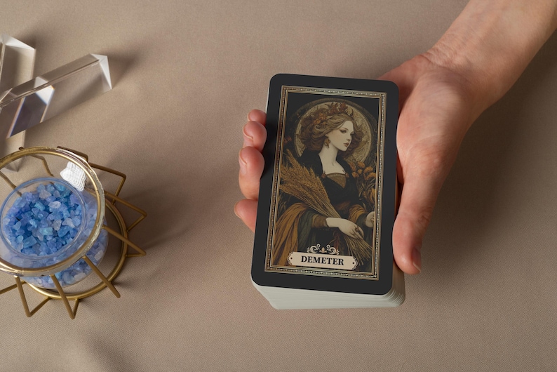 May include: A deck of tarot cards featuring a vintage illustration of Demeter, the Greek goddess of harvest. The card has a black border with gold accents and the name "Demeter" at the bottom. The deck is held in a hand.