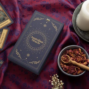 Peut inclure: Bo&icirc;te de jeu Anatolian Oracle bleu marine avec des lettres et des illustrations dor&eacute;es, entour&eacute;e de cartes de tarot, de bougies et d'un bol de p&eacute;tales de rose. Le texte "Anatolian Oracle" est visible.