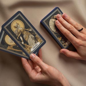 May include: A deck of tarot cards with navy blue borders and detailed illustrations. One card displays the word "THEMIS" and a depiction of a robed figure. The cards are held in a person's hands.