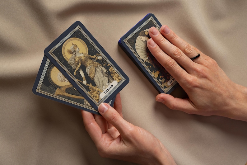 May include: A deck of tarot cards with navy blue borders and detailed illustrations. One card displays the word "THEMIS" and a depiction of a robed figure. The cards are held in a person's hands.