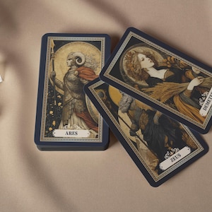 May include: A deck of tarot cards with navy blue borders, featuring illustrations of Greek gods and goddesses. The cards are stacked and fanned out, with the names "Ares", "Demeter", and "Zeus" visible. The artwork has a vintage aesthetic.