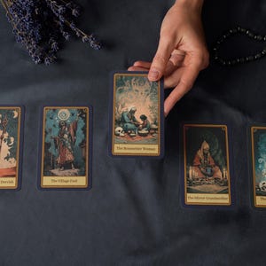 Peut inclure: Plusieurs cartes de tarot avec des illustrations d&eacute;taill&eacute;es et des titres, dispos&eacute;es sur un tissu bleu fonc&eacute;. Une main tient une carte, avec de la lavande s&eacute;ch&eacute;e et un collier de perles en arri&egrave;re-plan.
