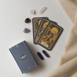 May include: A Mythology Oracle card deck with three cards showing Apollo playing a harp. The deck is navy blue with gold accents, and is surrounded by crystals in various colors. A beige cloth is in the background.